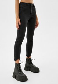Jeans ajustados de talle alto en negro con detalle de dobladillo deshilachado, combinados con botas negras de tacón grueso con cordones, que tienen una punta redondeada y suela gruesa.