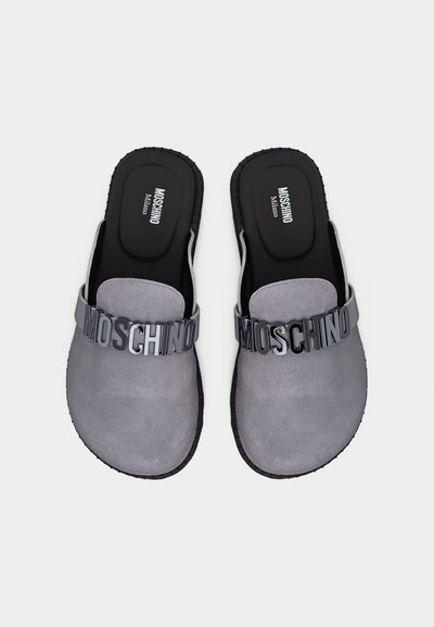 Γκρι σουέντ παντόφλες χωρίς κορδόνια με μαύρες σόλες και γράμματα "MOSCHINO" στα επάνω λουριά, μαύρα επενδυμένα πέλματα με την επωνυμία "Moschino Milano."