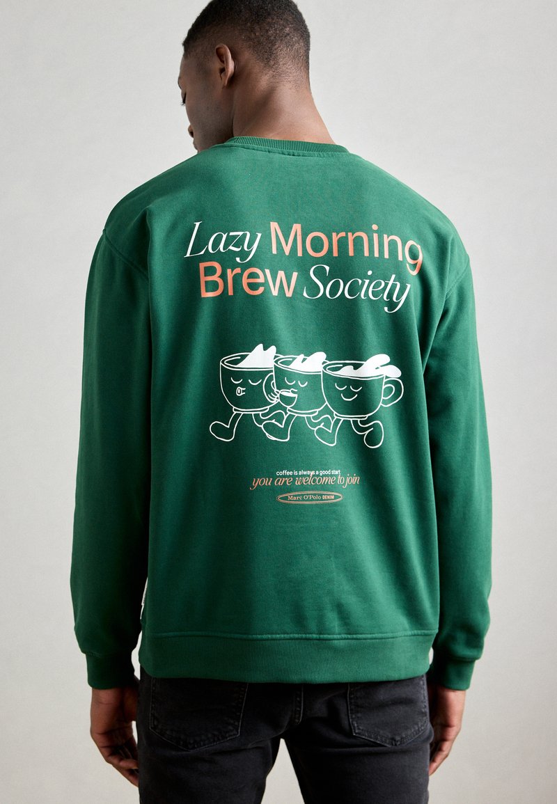 Sweatshirt vert avec du texte blanc et orange, des tasses à café en cartoon, et une coupe décontractée. Le design au dos met en avant des graphiques ludiques et un logo.