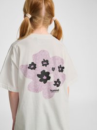 Kind mit roten Zöpfen, das ein weißes T-Shirt mit einer großen lila-schwarzen abstrakten Blumen-Grafik und dem "hummel"-Logo auf dem Rücken trägt.