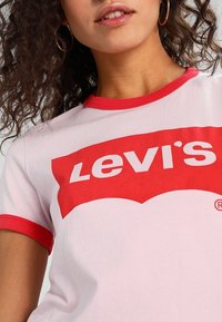 Rosa bomullströja med röda ringar vid ärmarna och en stor vit "Levi's"-logotyp på framsidan, med en klassisk crew neck-design.