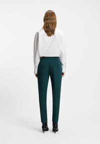 Weißes Hemd mit Kragen, kombiniert mit dunkelgrünen Hosen. Die Hose hat eine Gesäßtasche und ist im zeitlosen Schnitt gefertigt. Getragen mit schwarzen High Heels.