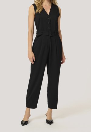 Frau trägt ärmellosen, schwarzen Jumpsuit mit Gürtel und Knöpfen sowie schwarze spitze Ballerinas, steht vor schlichtem Hintergrund.