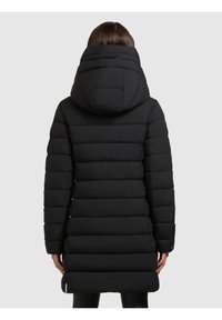 Person iført en lang sort polstret vinterjakke med en stor hætte, vist bagfra mod en neutral baggrund.