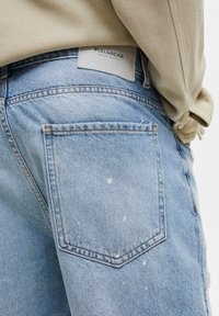 Vaaleansiniset farkut, joissa on kulunut pinta, näkyvät ompelu yksityiskohdat ja pieni valkoinen maalisirotus takataskussa. Brändin etiketti näkyvissä.