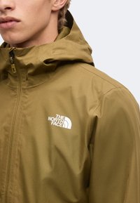 Personne portant une veste The North Face vert olive avec capuche et fermeture éclair à l'avant, épaule visible et visage partiel sur fond clair.