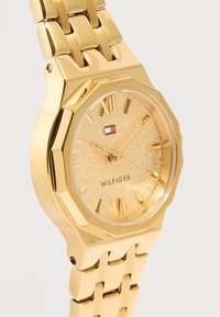 Ρολόι χειρός σε χρυσούς τόνους με υφή στο καντράν, ένδειξη ώρας με λατινικούς αριθμούς, τρία δείκτες και μπρασελέ με μεταλλικούς κρίκους με την επωνυμία "Hilfiger".