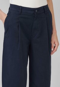 Pantaloni leggeri blu navy con plissettature frontali, tasche laterali e chiusura a bottone. Texture liscia, vestibilità su misura. Adatti per l'abbigliamento casual.
