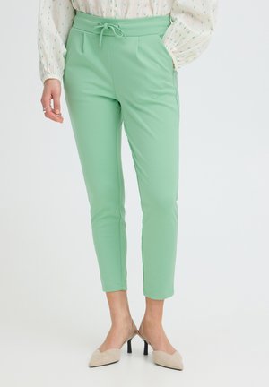 Femme portant un pantalon fuselé vert menthe avec poches latérales, associé à des talons beiges et un chemisier blanc à pois verts.