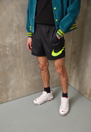 Mężczyzna w zielonkawoniebieskiej kurtce, czarnych szortach z neonowożółtym logo Nike, czarnych skarpetach i białych tenisówkach stojący przy szarej ścianie i podłodze.