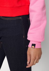 Personne portant un jean en denim bleu foncé et un sweat à blocs de couleurs avec un torse rouge et une manche rose, avec une étiquette logo sur le poignet.