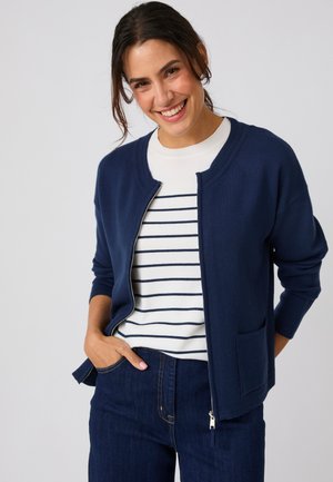 Mujer sonriente con cabello oscuro lleva un cárdigan azul marino con cremallera sobre una camisa blanca con rayas azul marino y vaqueros azul oscuro, posando con las manos en los bolsillos.