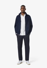 Marineblauwe ritsjack, gecombineerd met een lichtgestreept shirt, donkere broek en witte sneakers. Fijne textuur, casual fit en geen zichtbare logo's.