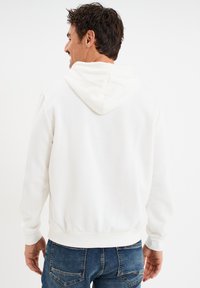 Sudadera blanca de algodón con capucha ajustable, puños y dobladillo acanalados. La vista trasera muestra una textura suave y un corte holgado. Se lleva con pantalones vaqueros azules.