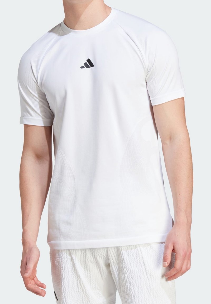 Muž v jednoduchém bílém tričku s krátkým rukávem sportovního střihu s malým černým logem Adidas na hrudi a texturovaných bílých šortkách.