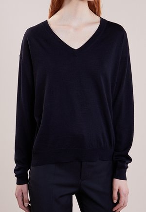 Woman wearing a loose black V-neck sweater and black pants, standing against a plain light background, hands relaxed by sides.