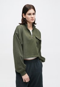 Νεαρή γυναίκα σε ελαιοπράσινο κοντό φούτερ με φερμουάρ και τσέπη με καπάκι, λευκό μπλουζάκι από κάτω και σκούρους παντελόνες με ελαστική μέση, μπροστά από ουδέτερο φόντο.