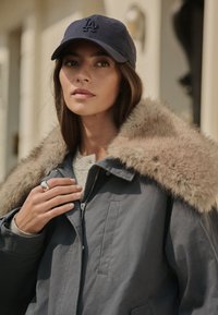 Veste extérieure grise avec un col en fausse fourrure, portée sur un pull de couleur claire. Une casquette marine avec un logo, et une bague décorative à la main.
