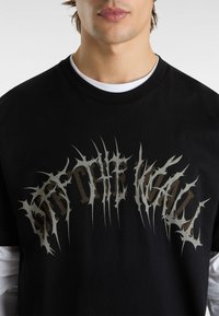 Sort bomulds T-shirt med en stor, tagget grafisk tekst "OFF THE WALL" i lys brun og grå. Rund halsudskæring og lagdelt hvid undertrøje synlig.