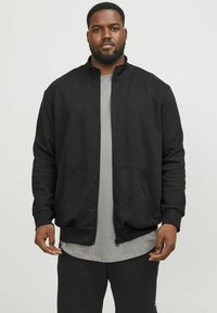 Chaqueta negra con cremallera y alto cuello, puños de canalé y bolsillos laterales, llevada sobre una camiseta gris. Textura suave, diseño casual.