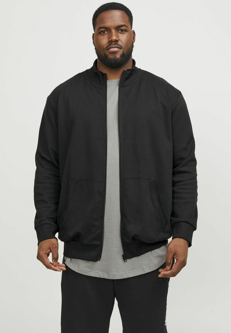 Chaqueta negra con cremallera y alto cuello, puños de canalé y bolsillos laterales, llevada sobre una camiseta gris. Textura suave, diseño casual.