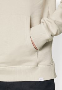 Sudadera beige con puños y dobladillo acanalados. Presenta un bolsillo canguro y una pequeña etiqueta en la parte inferior. Textura suave, diseño casual.
