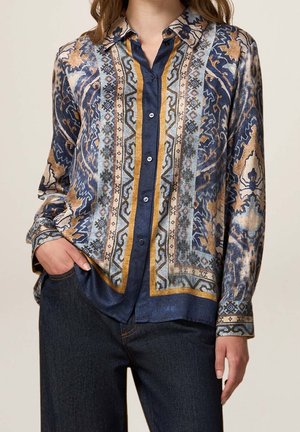 Femme portant une chemise à manches longues boutonnée avec des motifs géométriques et floraux complexes en bleu, beige et doré, associée à un jean foncé.