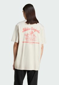 Grietai oversized t-shirt su rožine grafika ant nugaros, kuriame yra "Adidas Originals" ir sporto tema iliustracija. Trumpomis rankovėmis, lengvas prigludimas.