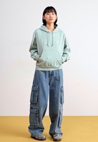 Sweat à capuche vert menthe avec poche avant et cordons de serrage ; associé à un pantalon cargo en denim bleu baggy avec de grandes poches latérales. Chaussures noires et blanches à rayures.