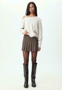 Sivi sv sweater bez ramena, smeđa mini suknja s nabranim detaljima i suptilnim prugama, crne čizme do koljena, s prozirnim najlonkama, na svijetloj pozadini.