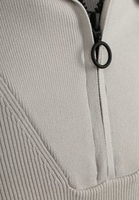 Lichtgrijs zip-up kledingstuk met ribbeltextuur, een hoge kraag en een zwarte ritsaccent met een ronde trekker.