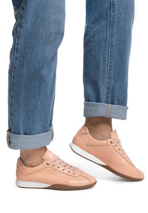 Osoba v modrých džínách s ohrnutými lemy a světle růžových teniskách na tkaničky, jedna noha mírně zvednutá, bílé pozadí.