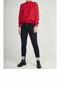 Rote Bluse mit langen Ärmeln und weißen Punkten, hochgeschlossen mit puffigen Schultern, kombiniert mit dunkelblauen high-waisted Jeans und schwarzen Combat-Stiefeln.