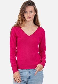 Fuchsia V-Ausschnitt Pullover aus einem weichen Strickstoff, mit langen Ärmeln und gerippten Bündchen, kombiniert mit hellblauen Jeans.