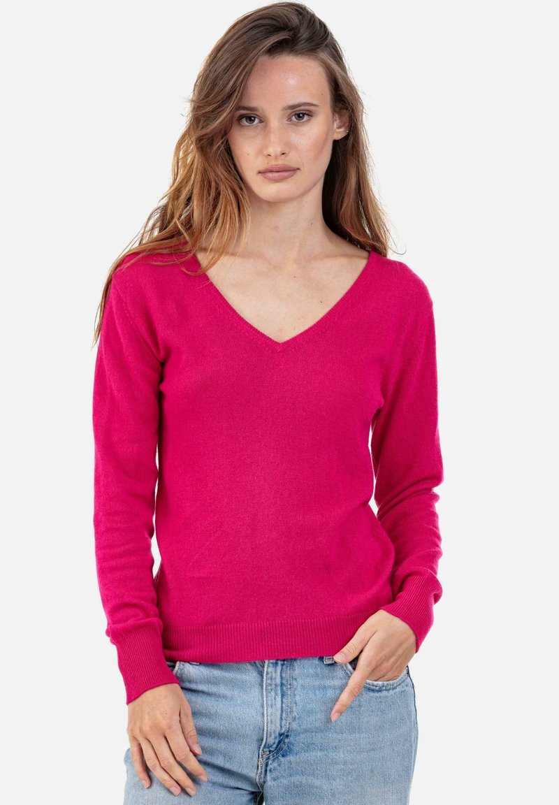 Fuchsia V-Ausschnitt Pullover aus einem weichen Strickstoff, mit langen Ärmeln und gerippten Bündchen, kombiniert mit hellblauen Jeans.