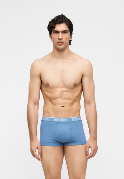 Homme torse nu aux cheveux courts et foncés portant un boxer Moschino bleu clair, debout devant un fond gris clair uni.