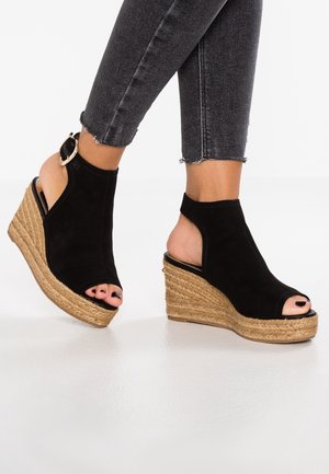 Voeten met zwarte open-toe sleehakken met zolen van juten touw en enkelbandjes, gecombineerd met cropped zwarte gewassen denim jeans.