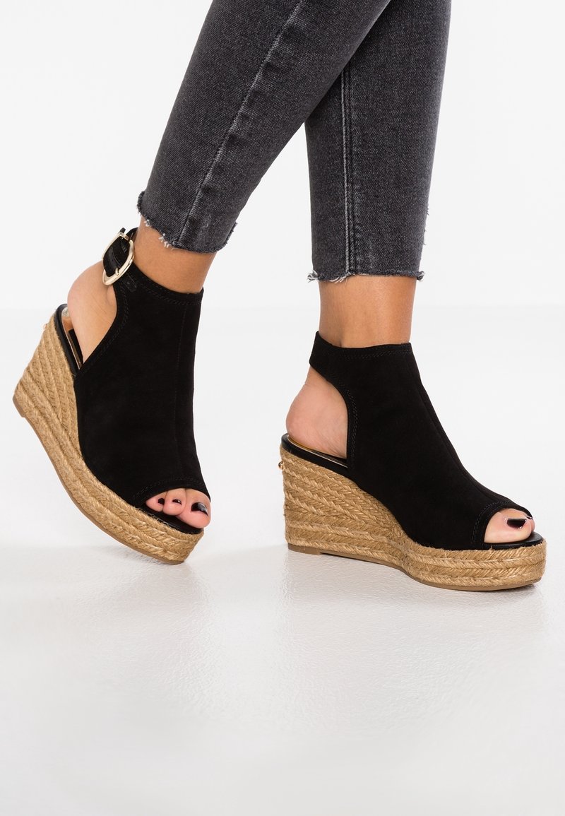 Pieds chaussés de sandales à talons compensés noires à bouts ouverts, semelles en corde de jute et brides à la cheville, associés à un jean court noir délavé.