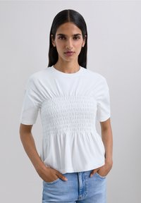 White short-sleeve shirt featuring smocking detail across the bust, relaxed fit, and a flared hem, paired with light blue jeans.