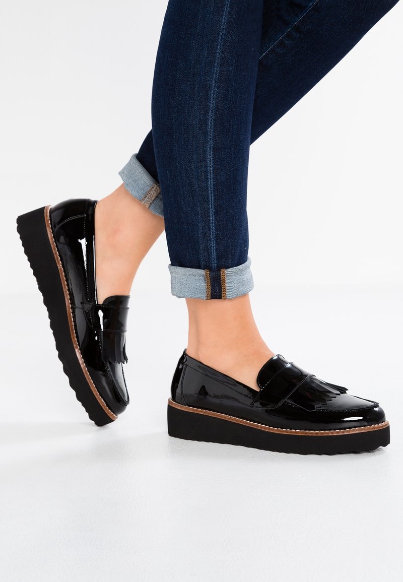 Mocassins à plateforme en cuir verni noir avec détail de franges portés avec un jean skinny bleu foncé retroussé sur fond blanc.