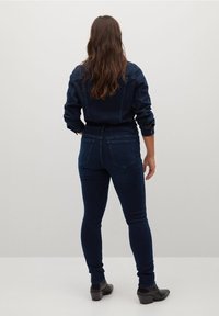 Mujer con cabello largo y castaño, vestida con chaqueta y jeans de mezclilla azul oscuro, de pie frente a una pared lisa de color gris claro.