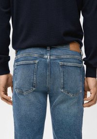 Tył osoby noszącej niebieskie dżinsy i ciemną koszulę z długim rękawem, z widocznymi dwoma tylnymi kieszeniami oraz brązową skórzaną łatą.