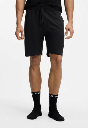 Mann trägt schwarze knielange Shorts mit Kordelzug und schwarze Socken und steht vor einem schlichten weißen Hintergrund.