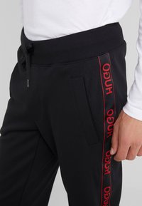 Schwarze Jogginghose mit elastischem Bund, verstellbarem Kordelzug, Seitentaschen und rotem Logodesign entlang des Beins. Weiche Stoffstruktur.