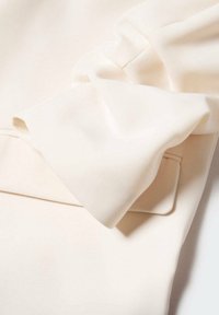Gros plan d'une manche en tissu écru avec un grand détail de nœud plié sur un rabat de poche rectangulaire.