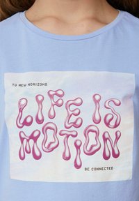 Lichtblauwe t-shirt met een bedrukt ontwerp met de tekst "LIFE IS MOTION" in roze, tegen een zachte, pastelachtergrond.