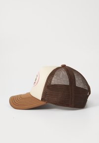 Ruskea ja beige trucker-lippis, jossa on verkko-osa takana, kaareva visori ja pyöreä logopatchi vaaleanpunaisena. Visorin ompeluominaisuudet.