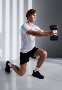 Muž v bielom športovom tričku s krátkym rukávom a čiernych šortkách vykonáva výpad s dvoma čiernymi činkami v minimalistickom prostredí.