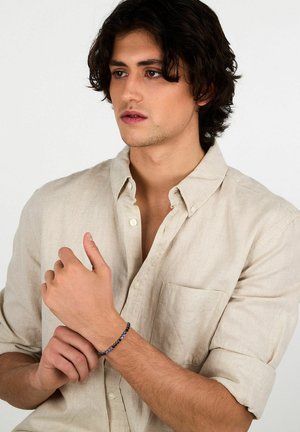 Braccialetto con perline di pietre blu scuro e chiaro su una fascia flessibile, indossato al polso, abbinato a una camicia di lino beige chiaro.