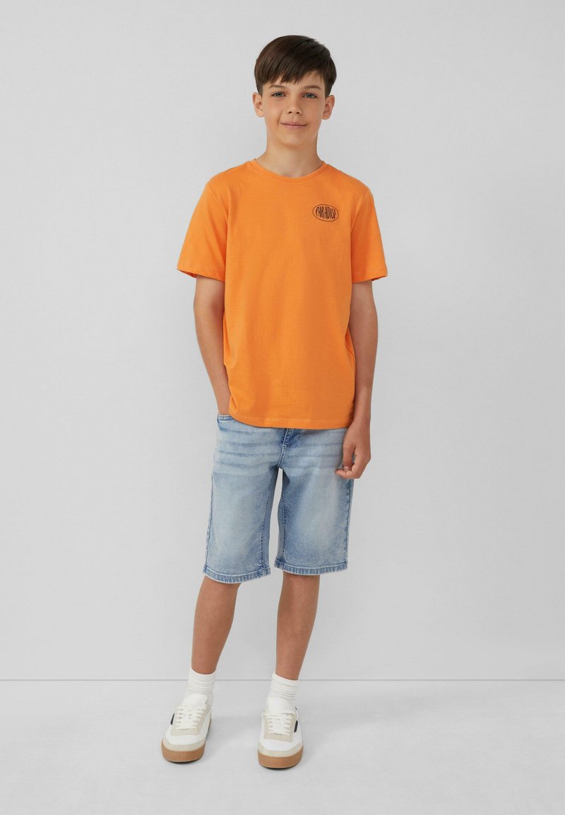 Oranje katoenen t-shirt met ronde hals en logo detail, gecombineerd met lichtblauwe denim shorts en witte sneakers met tan accenten.